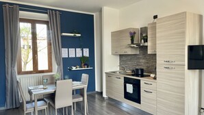 Une cuisine moderne avec des meubles de couleur bois clair, un four encastré et une cuisinière noire. On y trouve un coin repas avec une table et des chaises, ainsi qu’une fenêtre avec des rideaux.