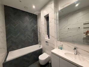 A modern bathroom with a herringbone-patterned wall, a white bathtub, a toilet, and a vanity with a mirror. - Casa Blu (Tbilisi)