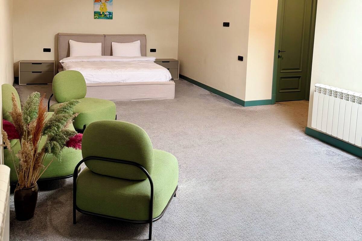 A hotel room with a bed, green chairs, a radiator, and a painting on the wall.