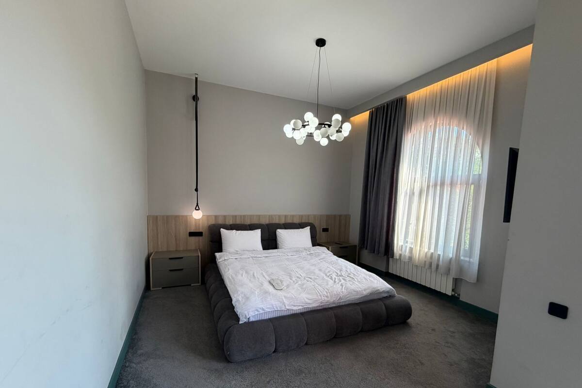 A modern bedroom with a large bed, a bedside table, a hanging light fixture, and a window with curtains.
