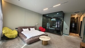 A modern bedroom with a large bed, a small round table, a yellow chair, and a glass-enclosed bathroom. - Casa Blu (Tbilisi)