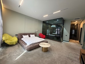 A modern bedroom with a large bed, a small round table, a yellow chair, and a glass-enclosed bathroom. - Casa Blu (Tbilisi)