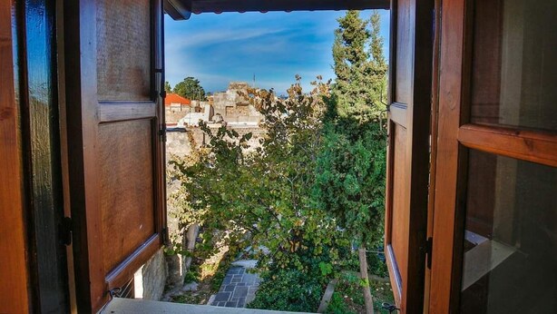A view from a balcony with a wooden railing, overlooking a courtyard with trees and a stone pathway. - Art House (Rhodes)