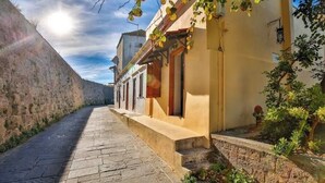 A narrow stone-paved street flanked by buildings with a stone wall on one side and a yellow building with a wooden door on the other. - Art House (Rhodes)