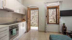 A compact kitchen with white cabinets, a stove, and a window with curtains. - Art House (Rhodes)