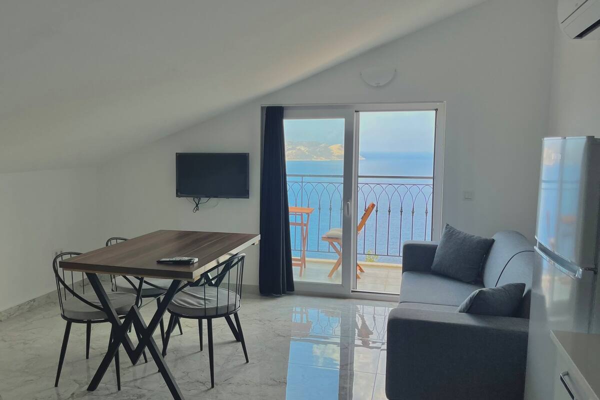 A modern apartment with a dining area, a sofa, a TV, and a balcony with a view of the sea.