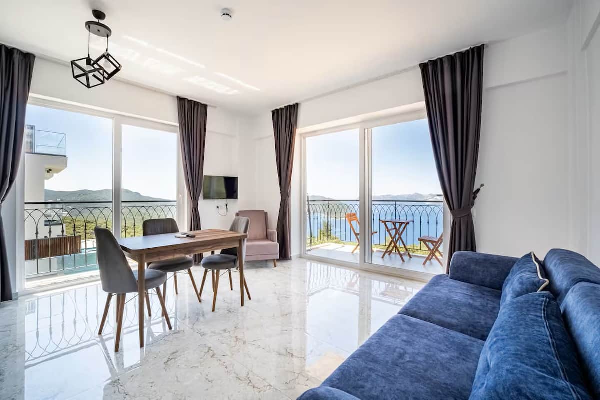 A modern living room with a large sofa, a dining table with chairs, and a balcony with a view of the sea.