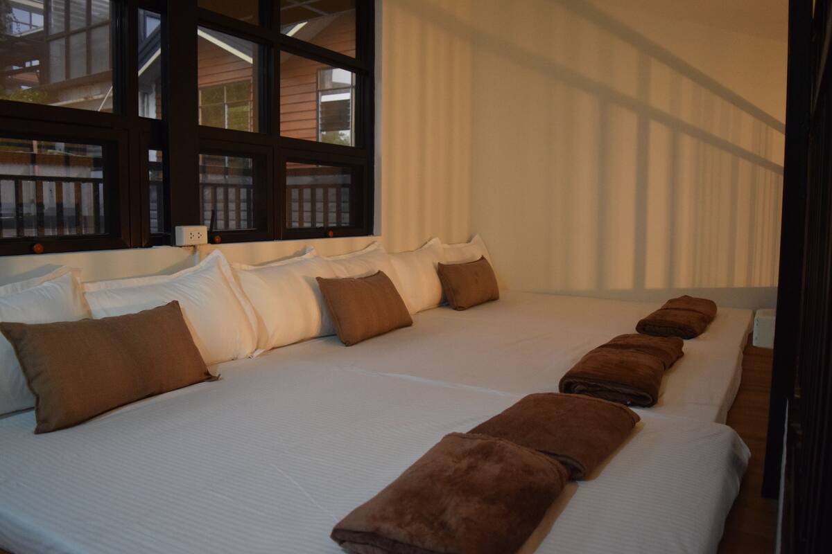 Una cama grande con muchas almohadas y toallas, una ventana con vista a una terraza de madera, y una cortina.