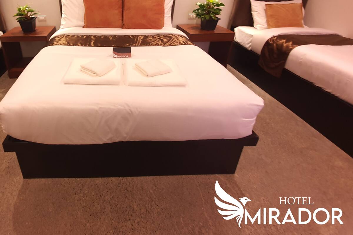 Habitación de hotel con una cama grande, dos mesitas de noche con lámparas, una planta en maceta y un letrero que indica el nombre del hotel.