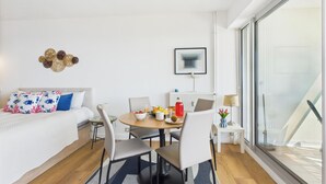 A modern, well-lit room with a bed, a dining table with chairs, a small side table with a lamp, and a large window offering a view.