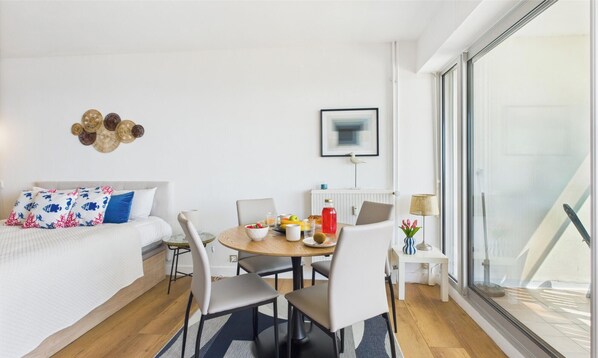 Une pièce moderne et bien éclairée, comprenant un lit, une table à manger avec des chaises, une petite table d’appoint avec une lampe, et une grande fenêtre offrant une vue dégagée.
