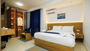 A hotel room with a large bed, a desk, a chair, a wardrobe, and an air conditioning unit.
