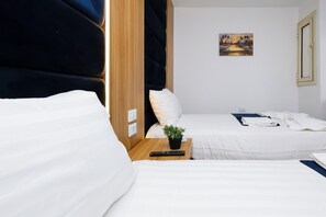 A hotel room with two single beds, a small table with a plant, a painting on the wall, and a door with a light switch.