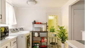 A compact kitchen with a washing machine, a coffee bar, and a glass table with a potted plant. - Family Home-Sleeps 8-Spacious 3BR-Free Parking-8 Min to PHL (Sharon Hill)