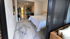 A modern hotel room with a marble floor, a bed with white bedding, a small kitchenette, and a view into the adjacent room.