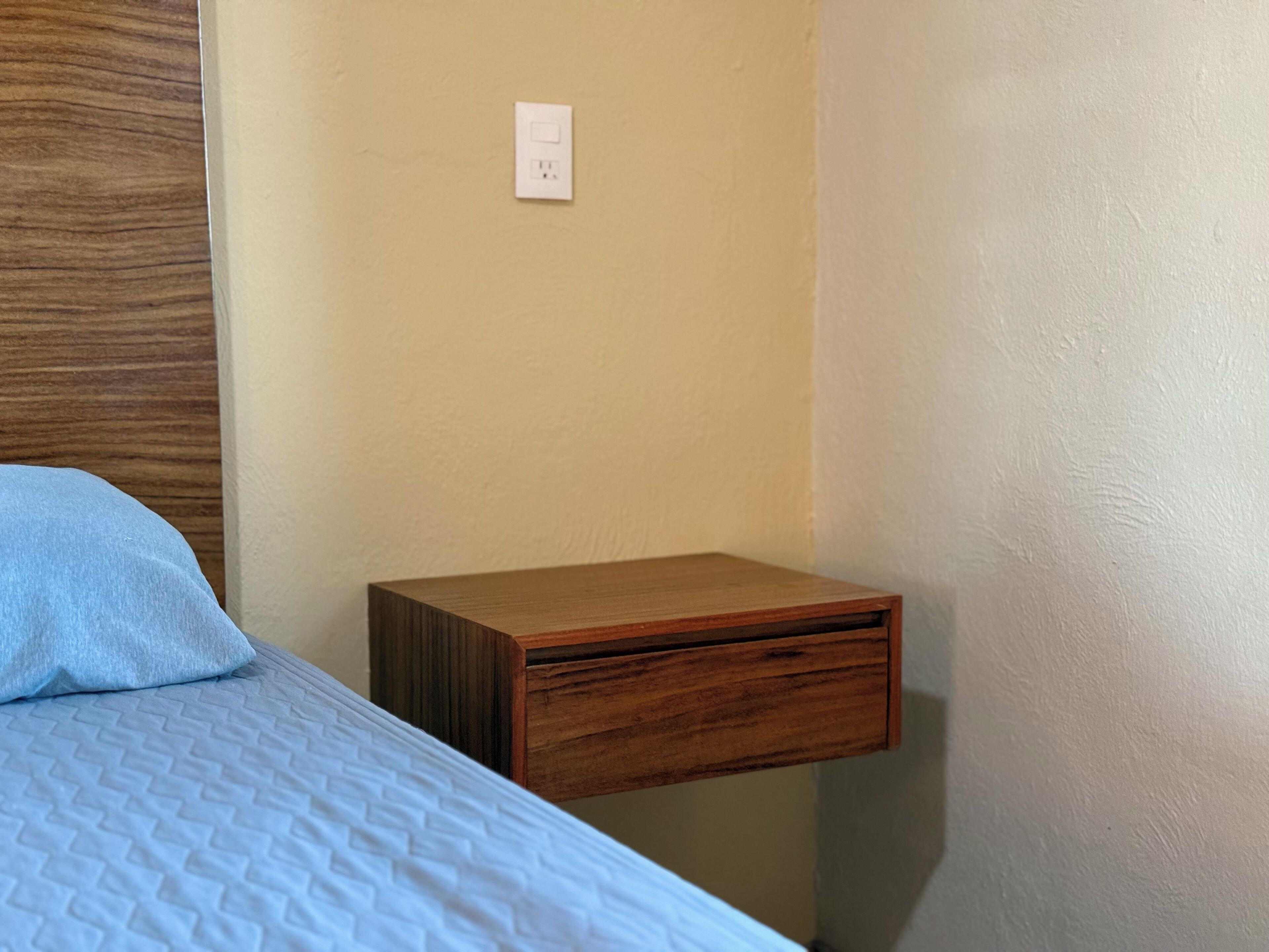 Un dormitorio con cama, una mesita de noche de madera y un enchufe.