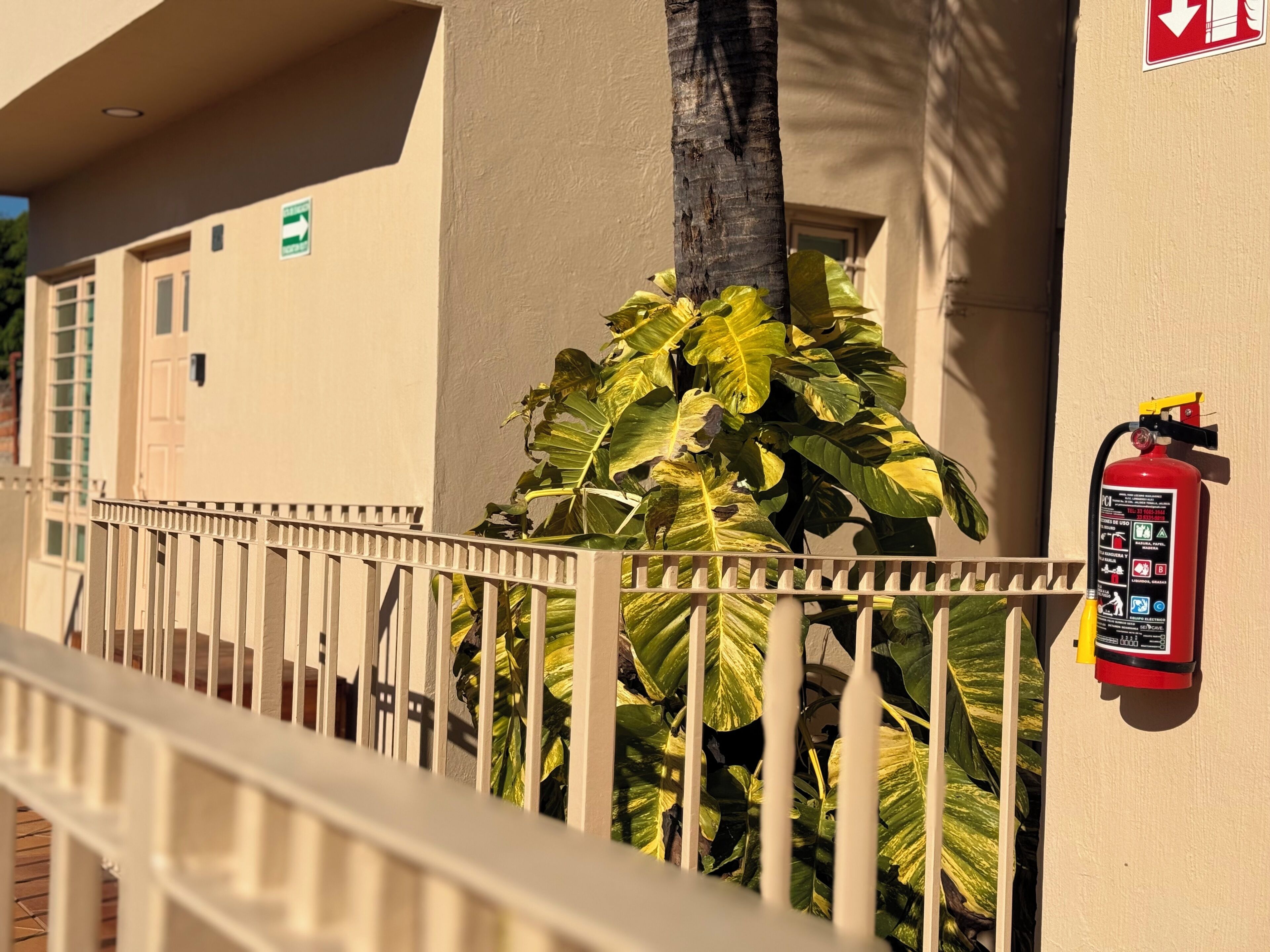 A fire extinguisher mounted on a wall, a white railing, and a building with a beige exterior and a door.