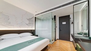 Une chambre d’hôtel moderne avec un lit, une cloison en verre, une salle de bain avec un miroir et un plancher en bois.