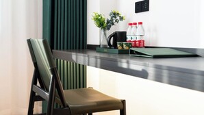 Une chambre d’hôtel moderne avec un bureau en bois foncé, une chaise verte, un vase avec des fleurs, une machine à café et deux bouteilles d’eau.