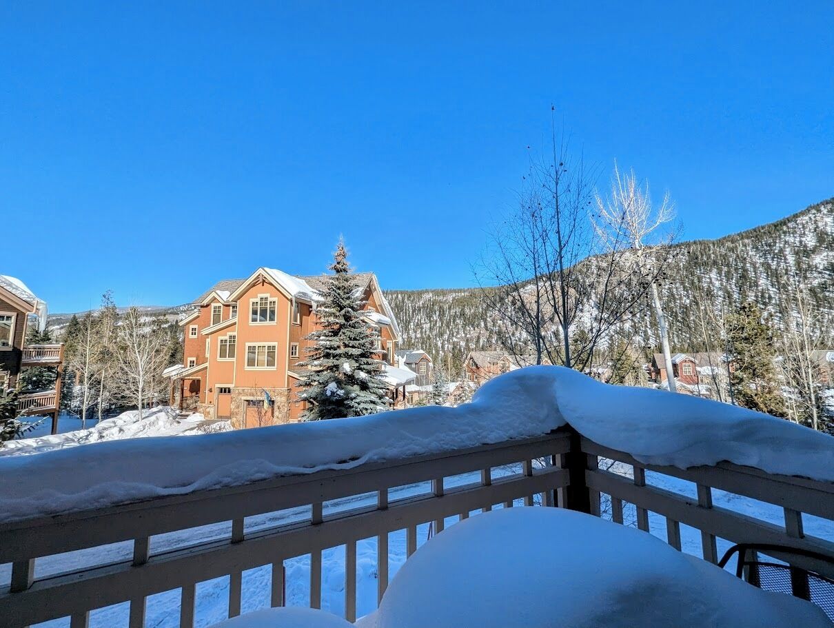 Una terraza de madera cubierta de nieve con vistas a un edificio grande de varios pisos, con fachada roja y techo nevado.