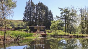 Eine Hütte mit einer Terrasse mit Blick auf einen Teich, umgeben von Bäumen und einer Wiese.