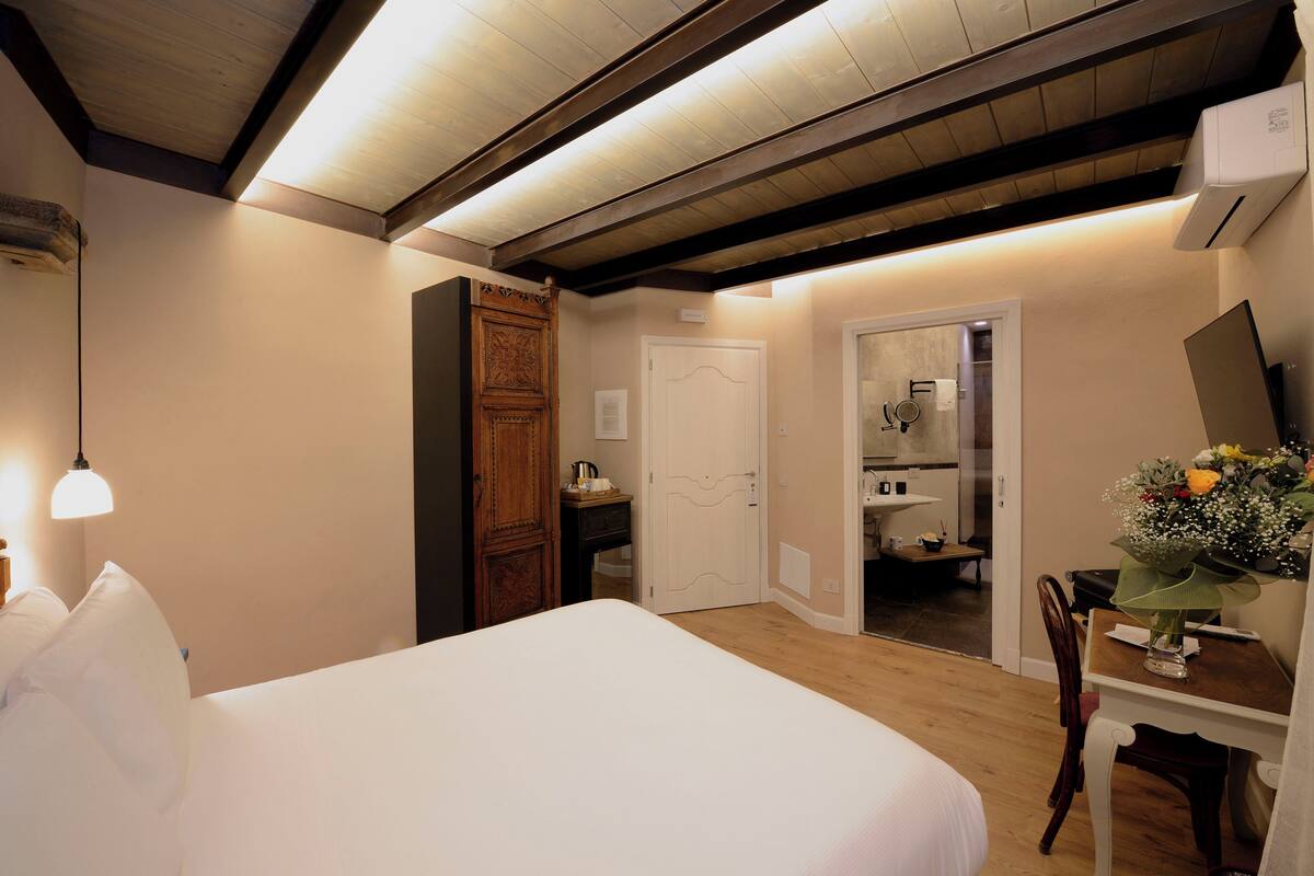 A bedroom with a large bed, a wooden wardrobe, a desk with a chair, and a bathroom visible through an open door.