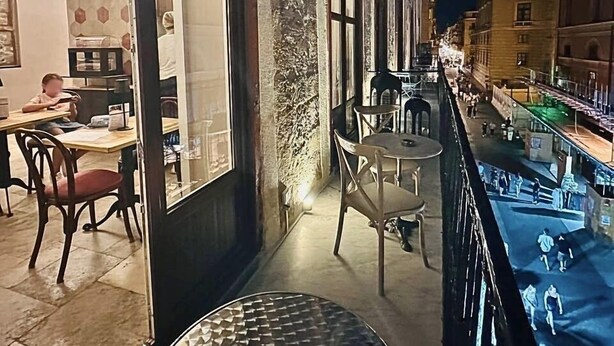 Un balcón con vista a una calle por la noche. Hay mesas y sillas en el balcón.