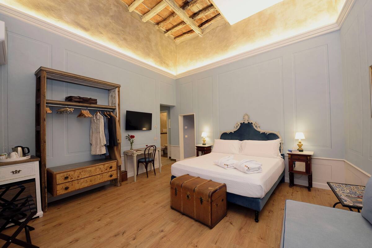 A hotel room with a large bed, a wooden wardrobe, a desk with a chair, and a small table.