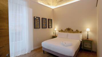 A neatly made bed in a room with a wooden floor, a window with sheer curtains, and two wall-mounted lamps.