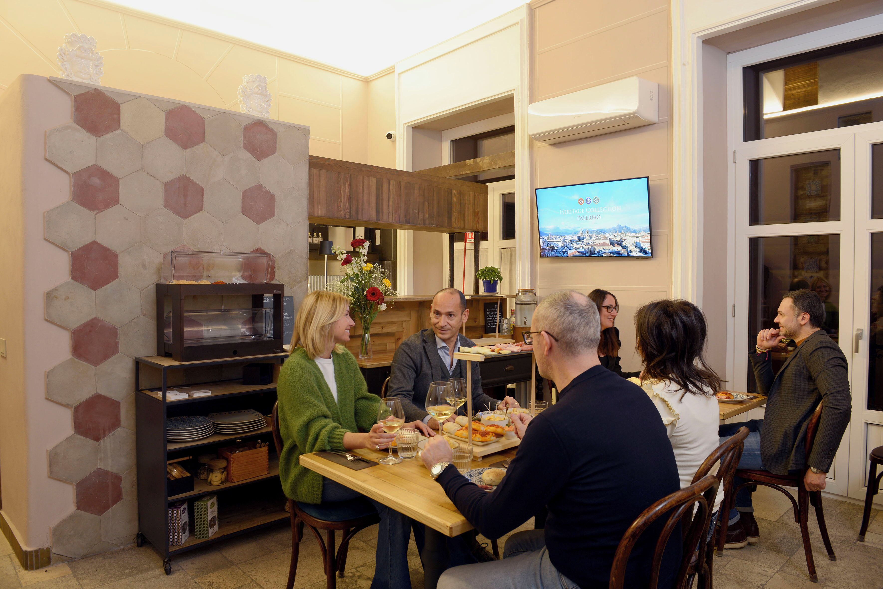 A group of people dining in a room with a hexagonal patterned wall, a television, and a wooden table.