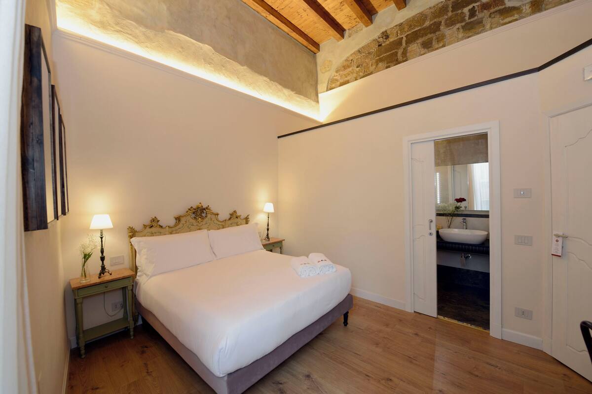 A bedroom with a bed, bedside lamps, a nightstand, a wooden floor, a brick wall, and a bathroom visible through an open door.