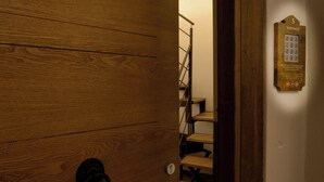 Una puerta de madera con un ojo de cerradura y un pomo metálico, que da acceso a una escalera con escalones de madera y barandilla metálica.