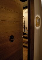 Una puerta de madera con un ojo de cerradura y un pomo metálico, que da acceso a una escalera con escalones de madera y barandilla metálica.