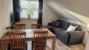 A cozy loft apartment with a dining area, a gray sofa with an anchor pillow, and a window with curtains. - Vacation home Küstennebel directly by the sea (Butjadingen)