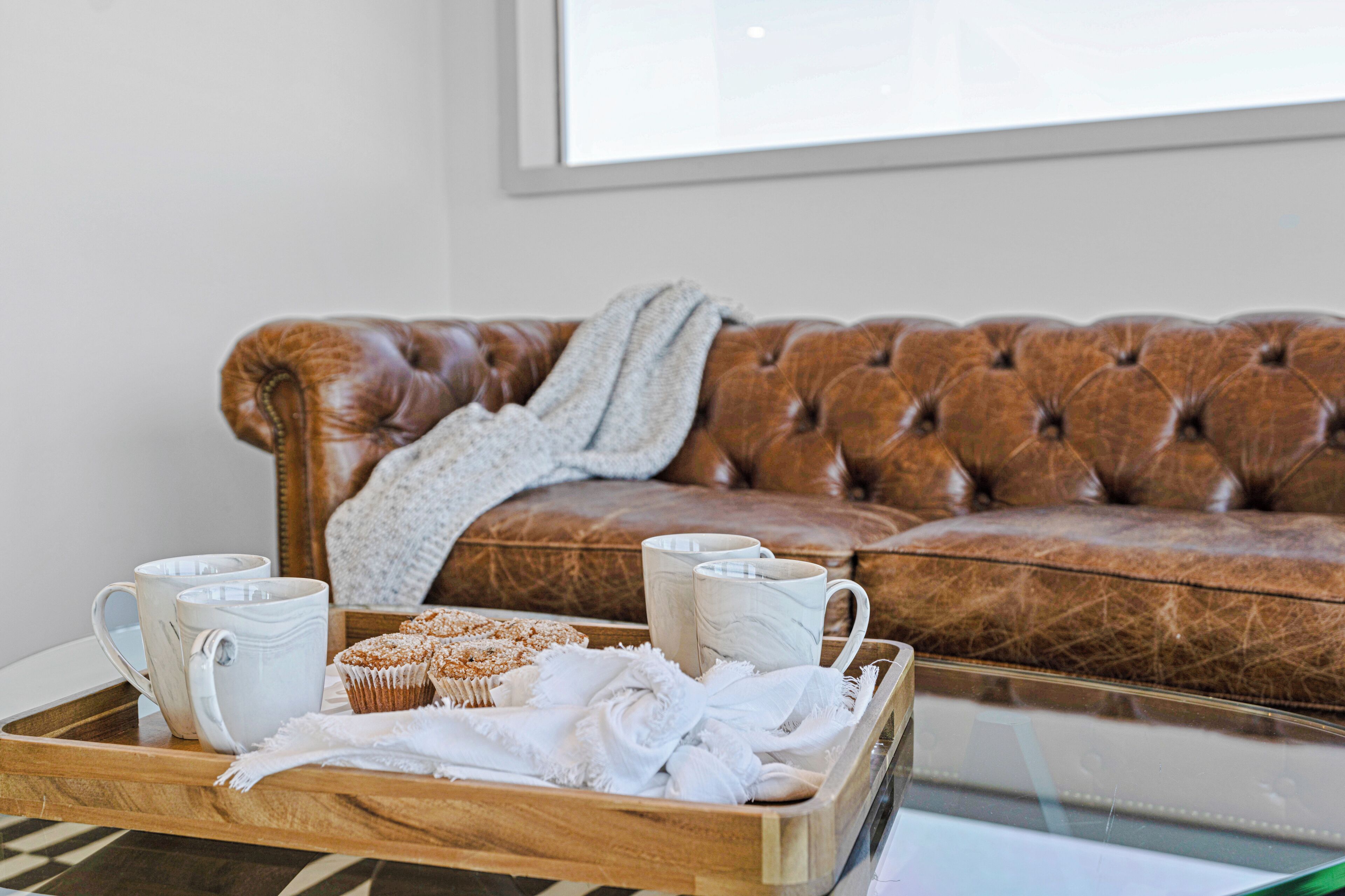 A wooden tray with white mugs, a muffin, and a white towel on a glass table in front of a brown leather sofa with a gray throw blanket.