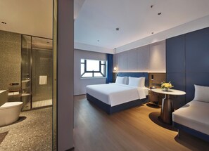 Una habitación de hotel moderna con ducha de vidrio, una cama grande con ropa de cama blanca, una mesita de noche con un jarrón de flores y una pequeña mesa redonda con una lámpara.