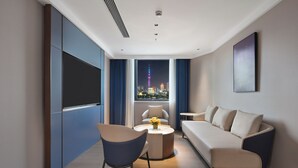 Ein modernes Hotelzimmer mit einer Couch, einem Sessel und einem runden Couchtisch. Ein großes Fenster bietet Blick auf die Stadt.