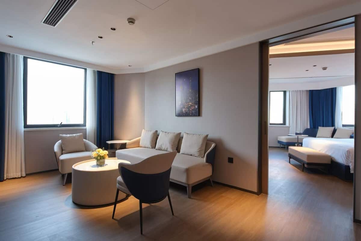 Una habitación de hotel moderna con un sofá, un sillón y una mesa redonda. Se ve una cama al fondo y un cuadro de un paisaje urbano en la pared.