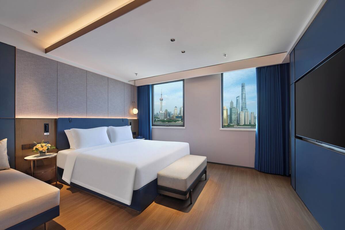 Una habitación de hotel moderna con una cama grande, un espacio para sentarse, una pequeña mesa con flores y vistas a la ciudad desde las ventanas.