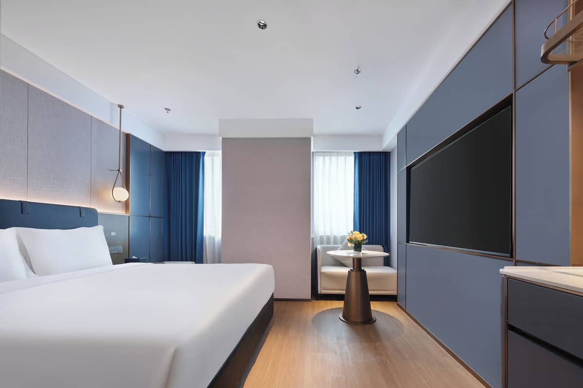 Una habitación de hotel moderna con una cama grande, una mesita de noche con un jarrón de flores, un rincón de descanso con una pequeña mesa y un televisor de pantalla plana grande montado en la pared.