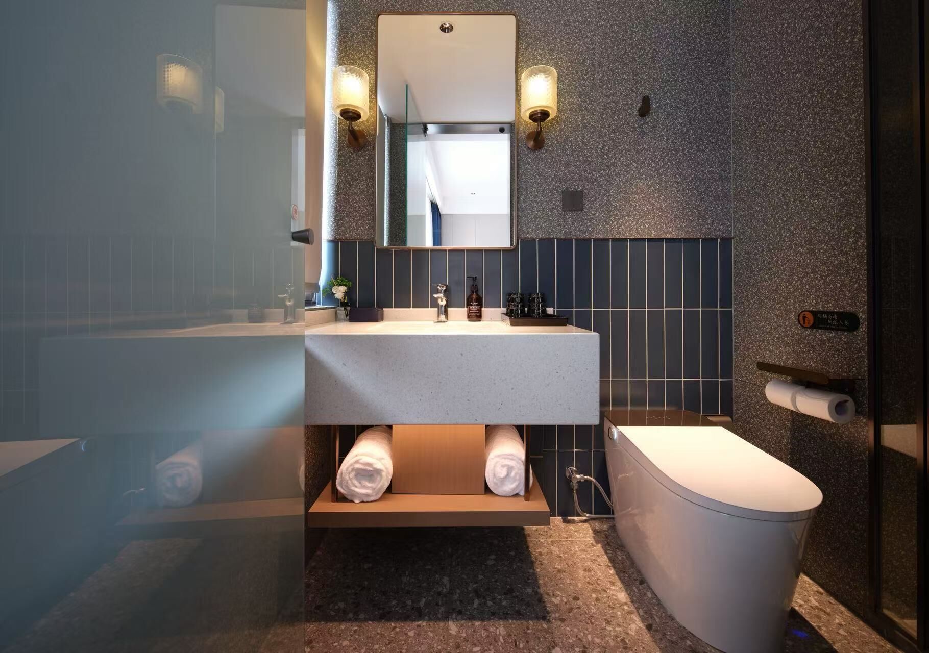 Een moderne badkamer met een witte wastafel, een spiegel erboven en een toilet. Handdoeken liggen op een plank en er is een douchecel zichtbaar.
