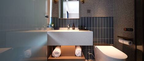 Een moderne badkamer met een witte wastafel, een spiegel erboven en een toilet. Handdoeken liggen op een plank en er is een douchecel zichtbaar.