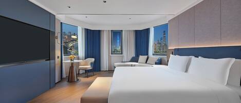 Una habitación de hotel moderna con una cama grande, una zona de estar con una mesita y vistas a la ciudad a través de amplios ventanales.