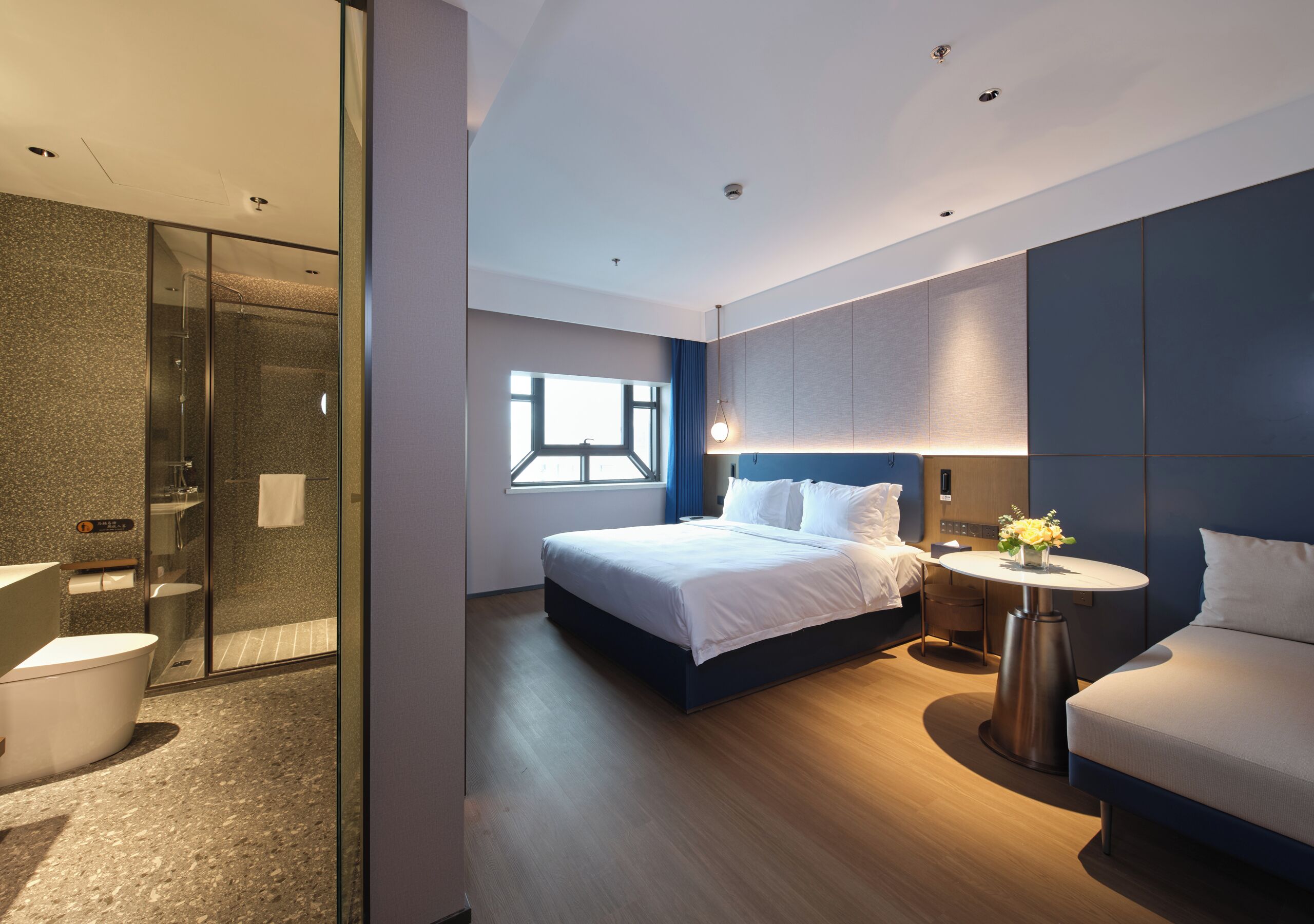 Una habitación de hotel moderna con ducha de vidrio, una cama grande con ropa de cama blanca, una mesita de noche con un florero y un sofá.