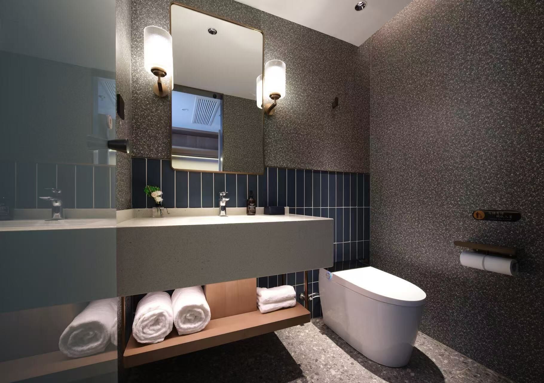A modern bathroom with a large mirror, two sinks, a toilet, and a shelf with towels.