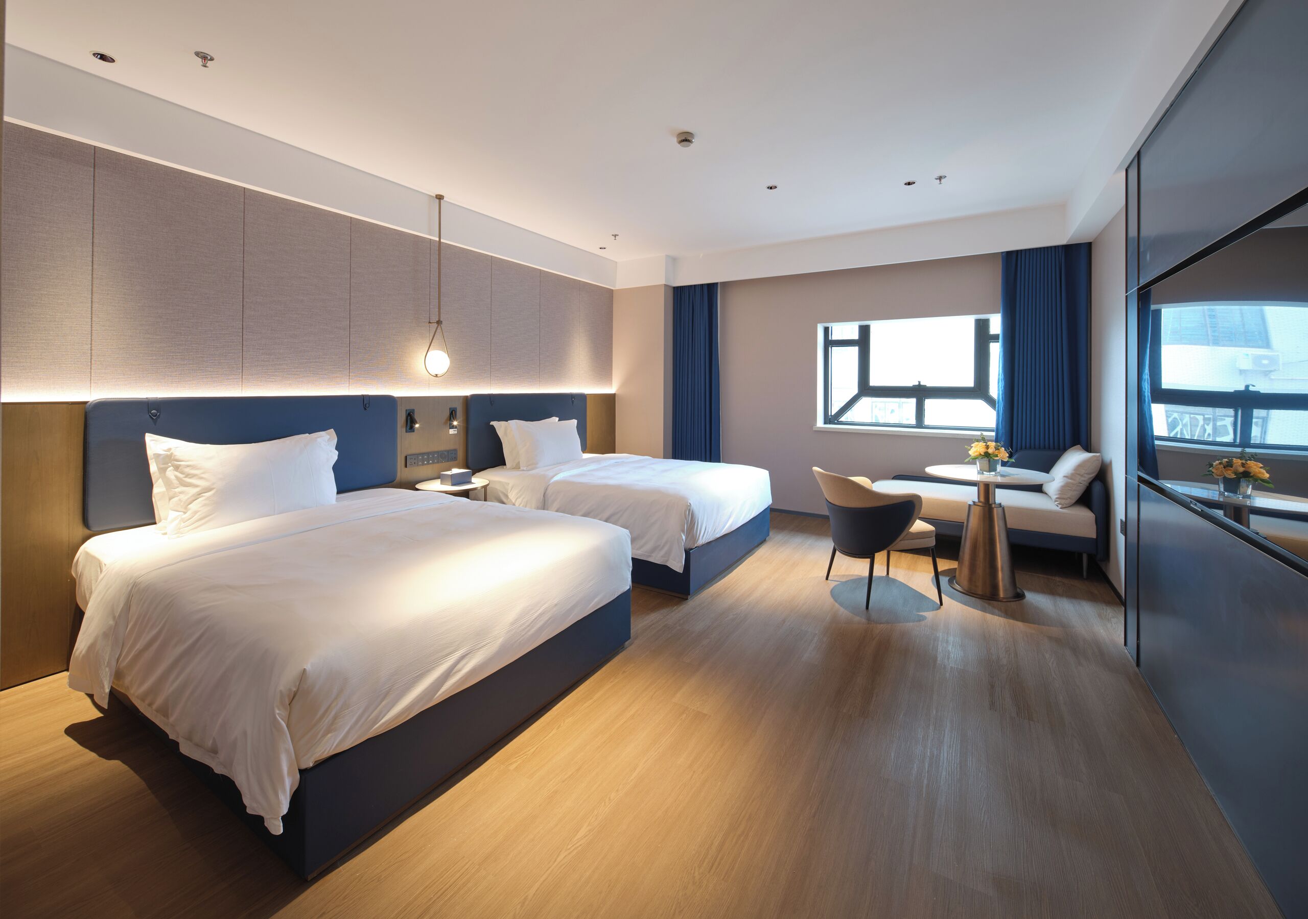 A modern hotel room with two beds, a small seating area, and a large window.