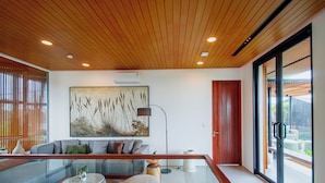 A modern living room with a wooden ceiling, a large abstract painting, a sofa set, a coffee table, and a glass railing overlooking a garden.