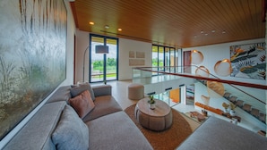 A modern living room with a large window, a sofa, a round coffee table, and a staircase with glass railings.