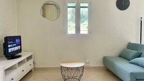 Une pièce avec un téléviseur posé sur un meuble blanc, un miroir rond accroché au mur, une fenêtre donnant sur un paysage verdoyant, une table ronde avec un plateau en bois et un canapé bleu.