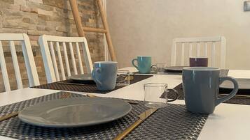 A dining table set with plates, cutlery, and blue and gray mugs, with a stone wall and a wooden ladder in the background.
